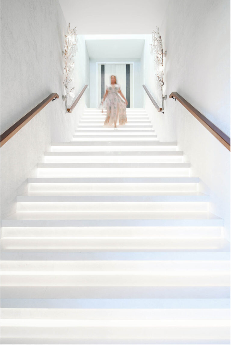 A person in a flowing, light-colored dress ascends a bright, illuminated staircase. The walls are white, and abstract sculptures are mounted on both sides. Handrails line the stairs, leading to a narrow archway at the top, creating a sense of elegance and modernity.