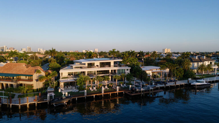 Aerial view of luxurious waterfront homes with large glass windows and lush gardens along a calm river, lined with docks and boats. The scene is bathed in late afternoon sunlight. In the distance, a skyline of a cityscape is visible against a clear blue sky.