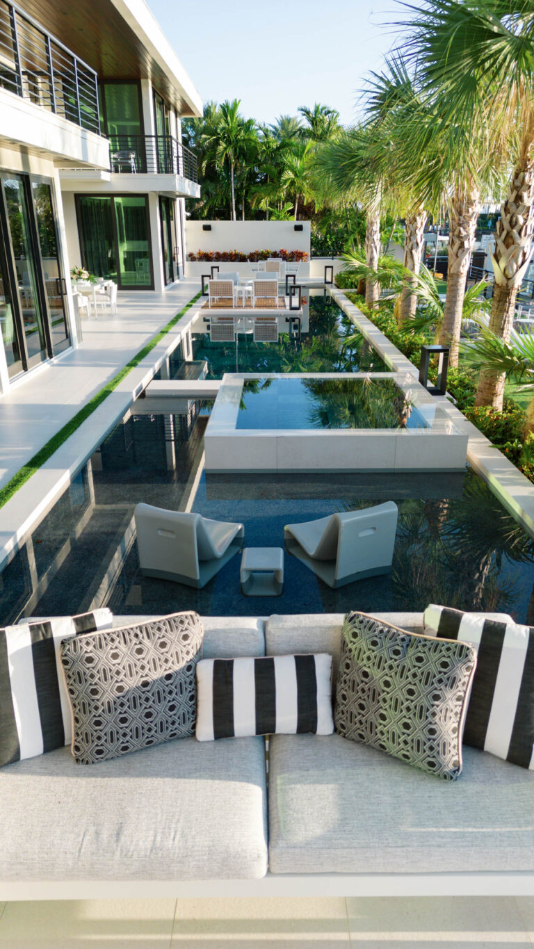 A modern outdoor patio features a sleek sofa with patterned and striped cushions overlooking a reflective pool. Two gray chairs are positioned by the water. The area is bordered by palm trees and a contemporary building with large windows, creating a luxurious, tropical ambiance.