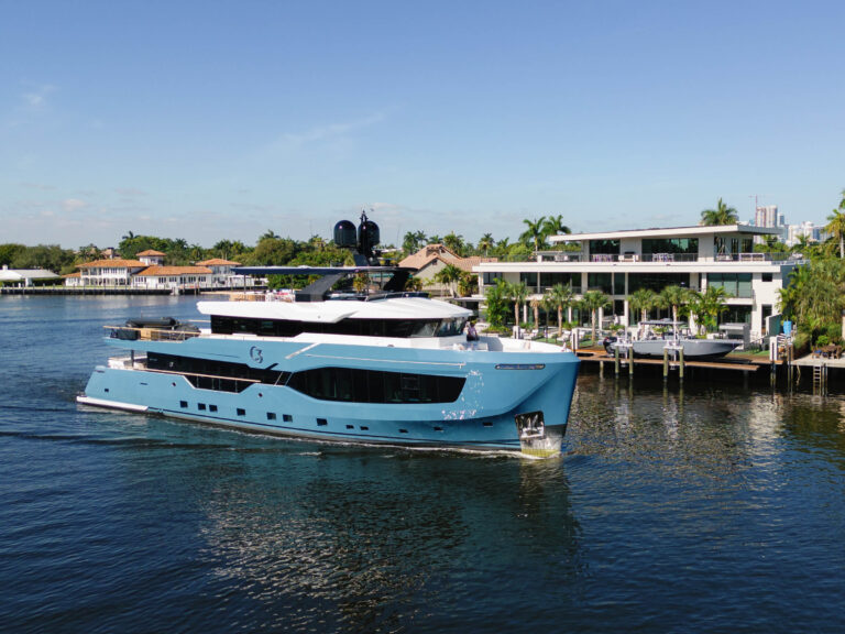 A large, sleek blue yacht cruises along a calm waterway under a clear blue sky. The vessel features multiple decks and modern design elements. In the background, luxurious waterfront homes and lush green palm trees line the shore, creating a scene of leisure and opulence.