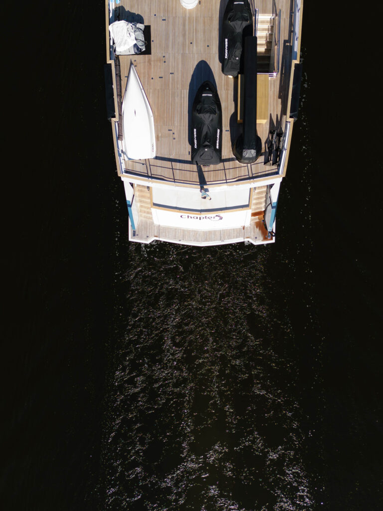 Aerial view of a boat on dark water. The deck has a white canoe, several covered objects, and visible wood planking. The rear of the boat has the word Chaplin and two small staircases leading to the water, creating a small wake behind the boat.