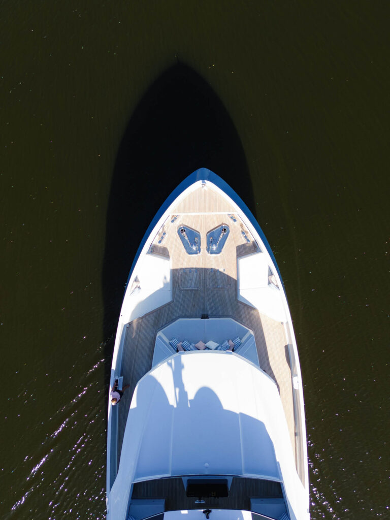 Aerial view of the bow of a white yacht on dark water, creating a prominent shadow. The deck features wood paneling, two seating areas, and visible railings. Sunlight reflects off the water, and part of the upper deck structure is visible at the bottom of the image.
