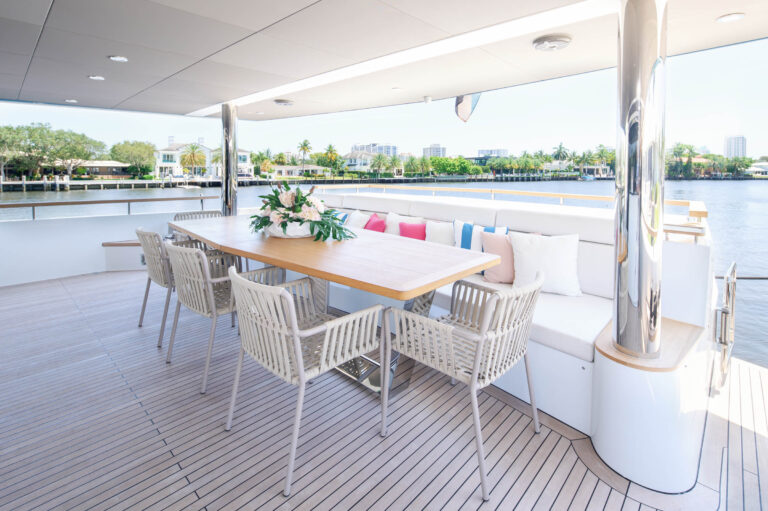Spacious yacht deck with a wooden dining table surrounded by white wicker chairs and a cushioned bench. The table features a floral centerpiece. The deck overlooks a tranquil body of water with buildings and palm trees in the distance under a clear blue sky.