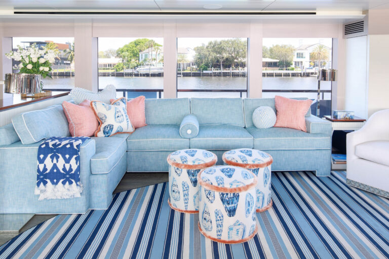 A living room with a light blue L-shaped sofa adorned with pink and patterned pillows. Two cylindrical stools with blue and white designs serve as a coffee table on a blue-striped rug. Large windows offer a view of water and trees, with a white armchair on the right.
