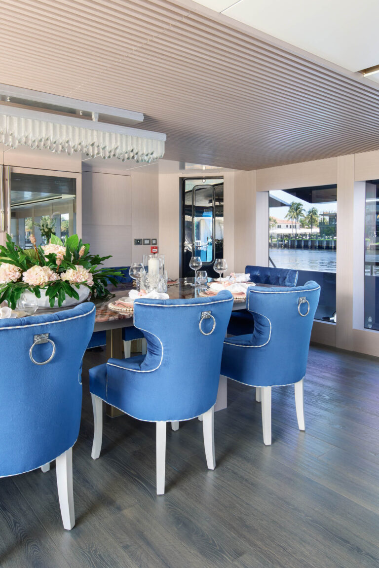 Elegant dining room with a polished wooden table set with fine china, crystal glassware, and floral centerpieces. Blue upholstered chairs with ring pull accents surround the table. Large windows offer a view of water and greenery, while a modern chandelier hangs from the ceiling.