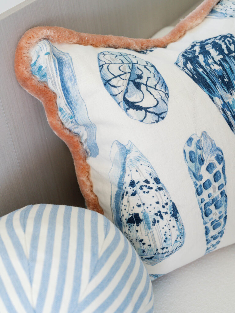 Close-up of two decorative pillows on a couch. The main pillow features blue seashell illustrations with a fuzzy orange trim. The companion pillow has a light blue and white diagonal stripe pattern. The backdrop is a neutral-toned wall or couch fabric.