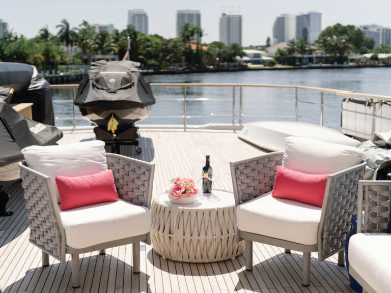 Two wicker chairs with white cushions and pink throw pillows face each other on a yacht deck. Between them is a round table with a bottle of wine and two glasses. The background shows a calm river, lush greenery, and a city skyline with tall buildings under a clear blue sky.