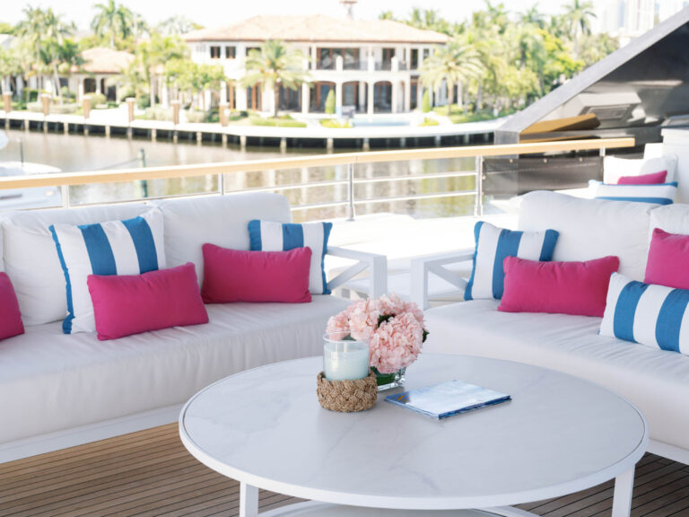 Luxury yacht deck with white cushioned seating featuring blue-striped and pink pillows. A round white table in the center displays a blue glass candle, pink hydrangeas, and a magazine. In the background, waterfront homes and lush palm trees are visible across the water.