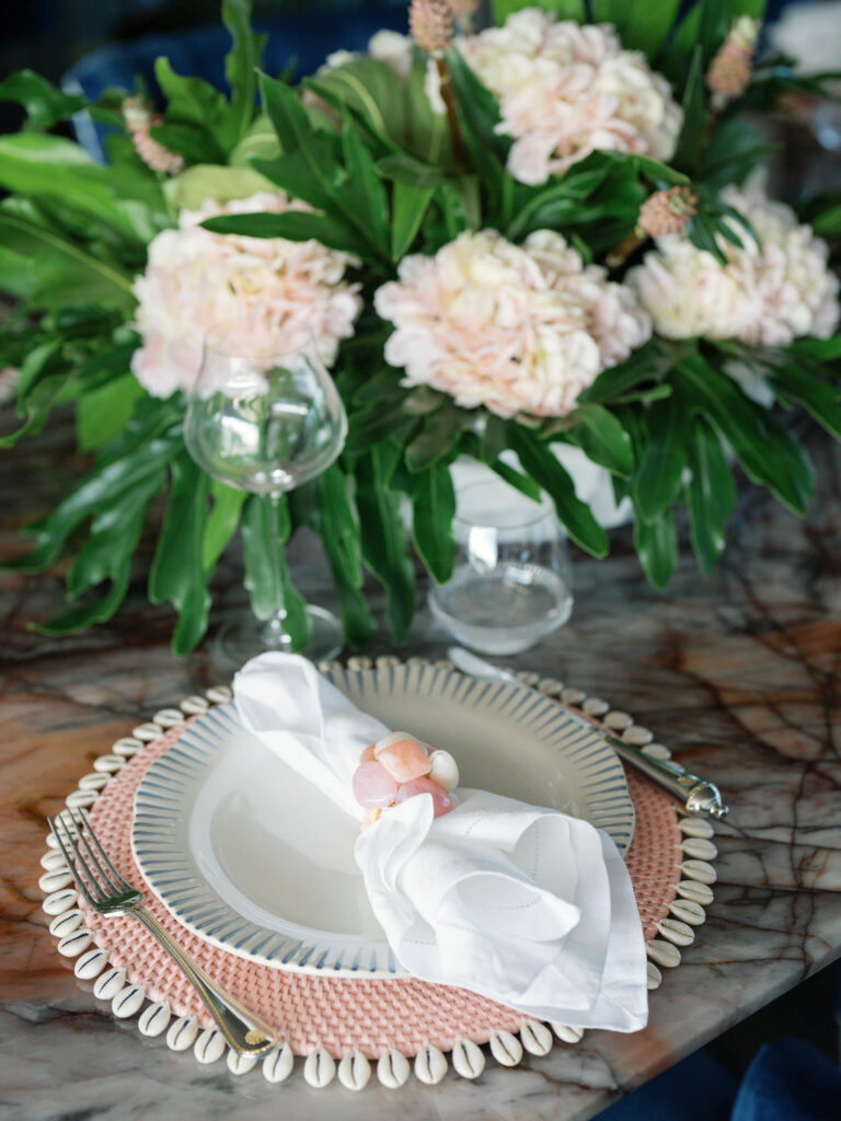 A table setting with a plate on a pink, beaded placemat adorned with white shell accents. A white napkin with a pink flower napkin ring rests on the plate. Beside the plate is a fork and knife. A wine glass is partially filled, and theres a background arrangement of pale pink flowers and green leaves.