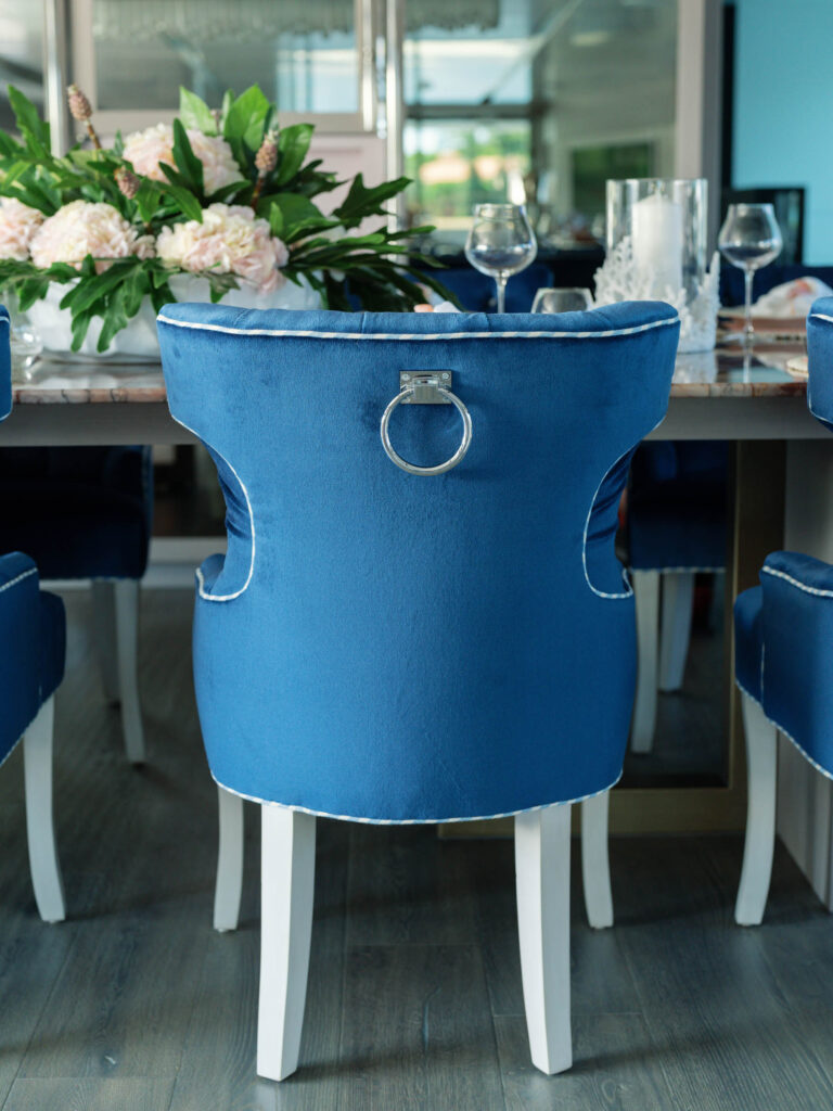 A stylish dining chair with rich blue velvet upholstery, featuring a silver ring handle on the back, is positioned at a dining table. The chair has white legs and sits on a wood floor. The table is adorned with a floral arrangement and glassware, reflecting an elegant setting.