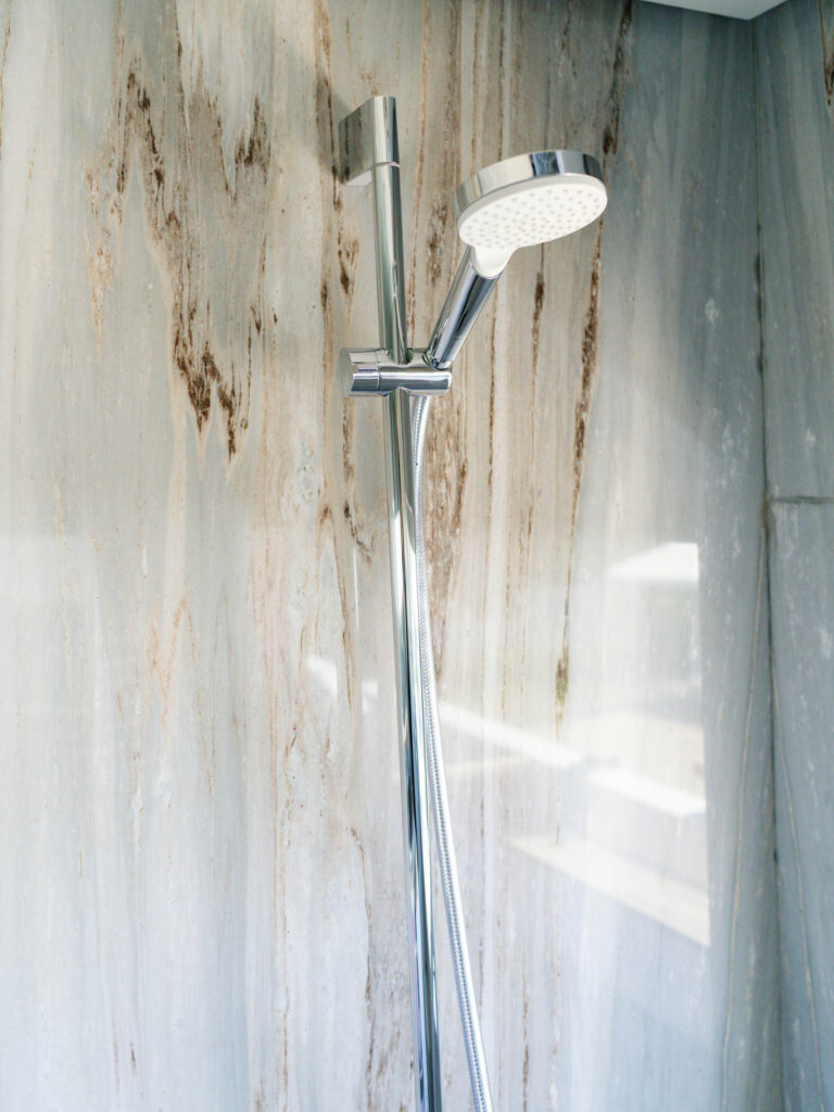 A modern shower head with a sleek chrome finish is mounted on a vertical rail against a marble-patterned wall. Sunlight streams in from the right, casting soft reflections on the wall and the metal surface, emphasizing the clean and minimalist design of the bathroom.