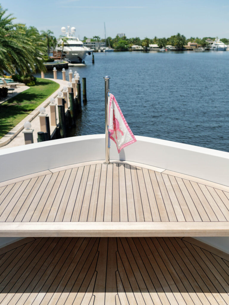 View from the bow of a yacht showing a wooden deck with a pink-bordered flag on a pole. The yacht is docked near a waterfront lined with palm trees and luxury homes. Another yacht is visible in the background under a clear blue sky.