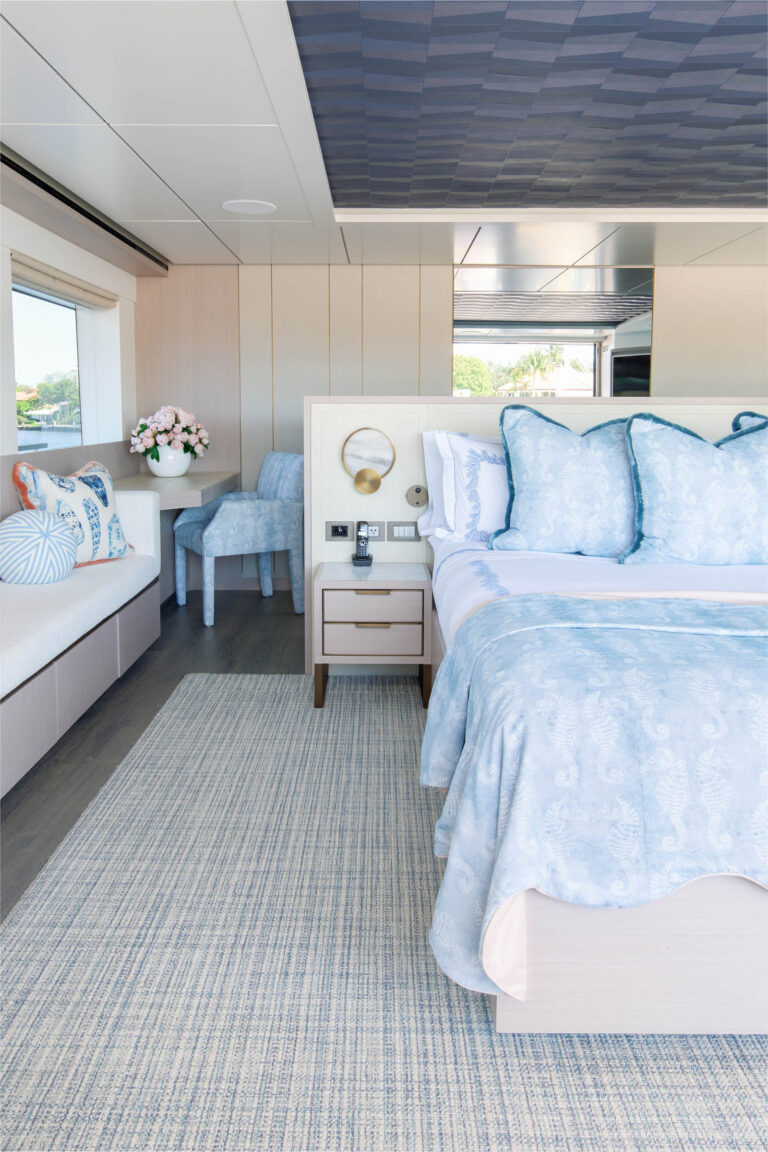 A bright bedroom features a bed with blue patterned bedding and multiple pillows. A small bedside table with a lamp is beside the bed. Theres a window seat with cushions and a flower arrangement. Two upholstered chairs sit at a small table. The room has light wood and elegant decor.