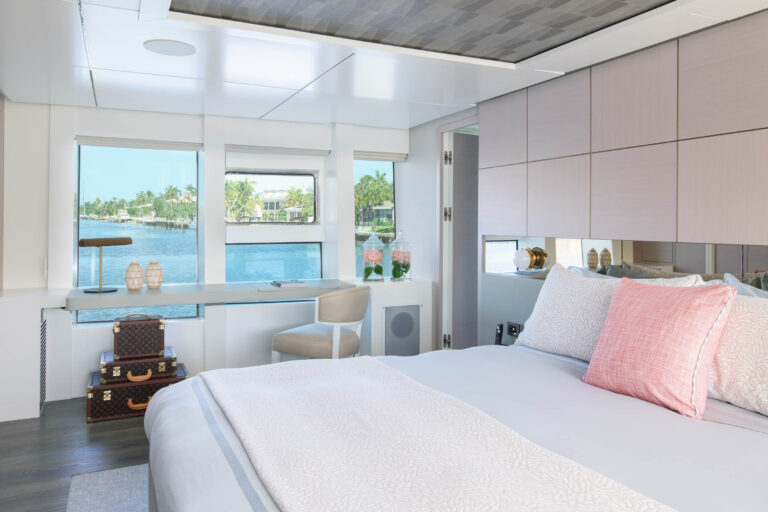 A serene bedroom scene features a large bed with white linens and a pink pillow. Theres a stack of decorative suitcases on the floor. The room has large windows offering a view of the water and greenery outside. A white chair and small desk with decor are positioned by the windows.