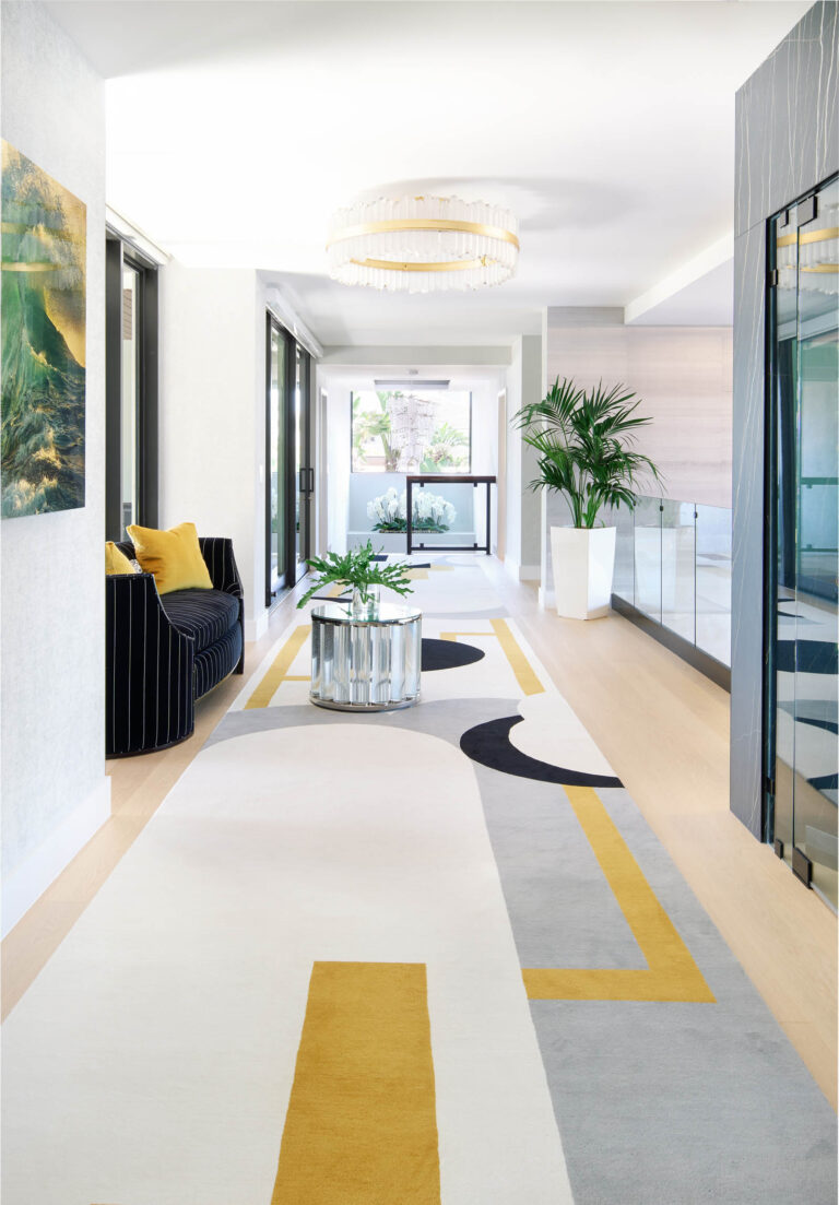 A modern, airy hallway with a geometric carpet in white, gray, black, and mustard. A black striped armchair with yellow cushions sits beside a glass wall. A metallic coffee table holds a plant, while a large potted plant is near the glass railings. A circular chandelier hangs above.