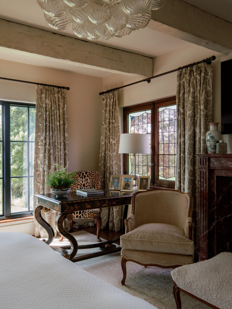 A cozy corner of the exuberant interior of this French Norman-inspired home features a dark wood desk with a leopard-print chair, framed photos, and a potted plant. Floral curtains frame tall windows beneath exposed beams, and a white bed is partly visible.
