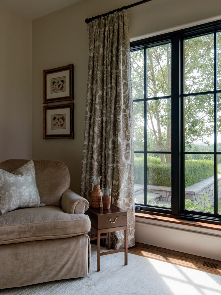 A cozy beige armchair with a patterned pillow sits by a window with leafy drapes. A wooden side table holds decorative vases. Outside the black-framed window, greenery and trees are visible. Two framed artworks hang on the light wall above the chair.