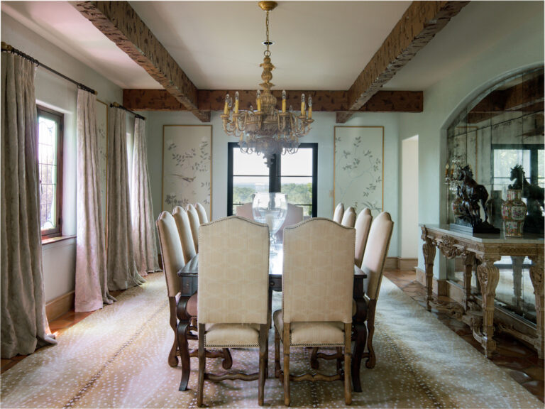 Elegant dining room with a long dark wood table, twelve cream upholstered chairs, beige patterned rug, ornate chandelier, exposed wooden ceiling beams, large windows with sheer curtains, and a sideboard with decorative statues and a mirror.