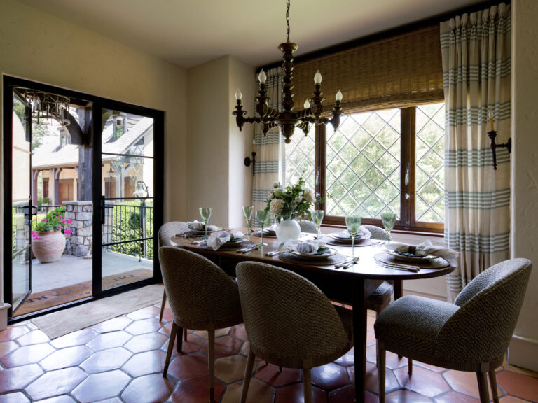 A cozy dining room in the exuberant interior of this French Norman-inspired home features six upholstered chairs around a dark wood table, vintage chandelier, and large plaid-curtained windows overlooking greenery and a patio with a potted plant.