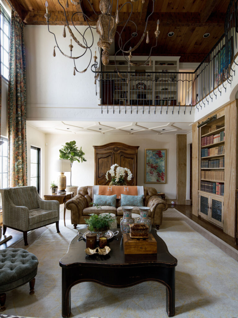 Elegant living room with high ceilings, chandelier, tan leather sofa, armchairs, large wooden coffee table with decor, built-in bookshelves, tall windows with patterned curtains, potted plants, and a loft area with black railing above.