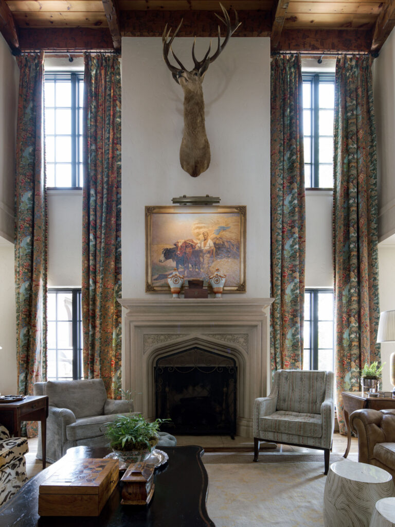 A grand living room with tall floral drapes flanking two windows, a stone fireplace mantel, a painting and mounted deer head above it, two armchairs, decorative objects, and a dark wood coffee table with a plant and boxes. Exposed wood beams line the ceiling.
