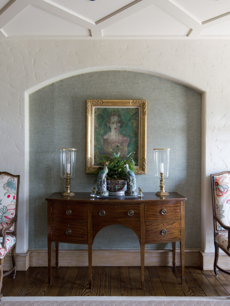 A wooden console table with brass handles sits against a wall niche with blue wallpaper. On the table are two glass hurricane candle holders, two porcelain peacock figurines, and a potted plant. Above hangs a framed portrait of a woman. Two chairs flank the table.