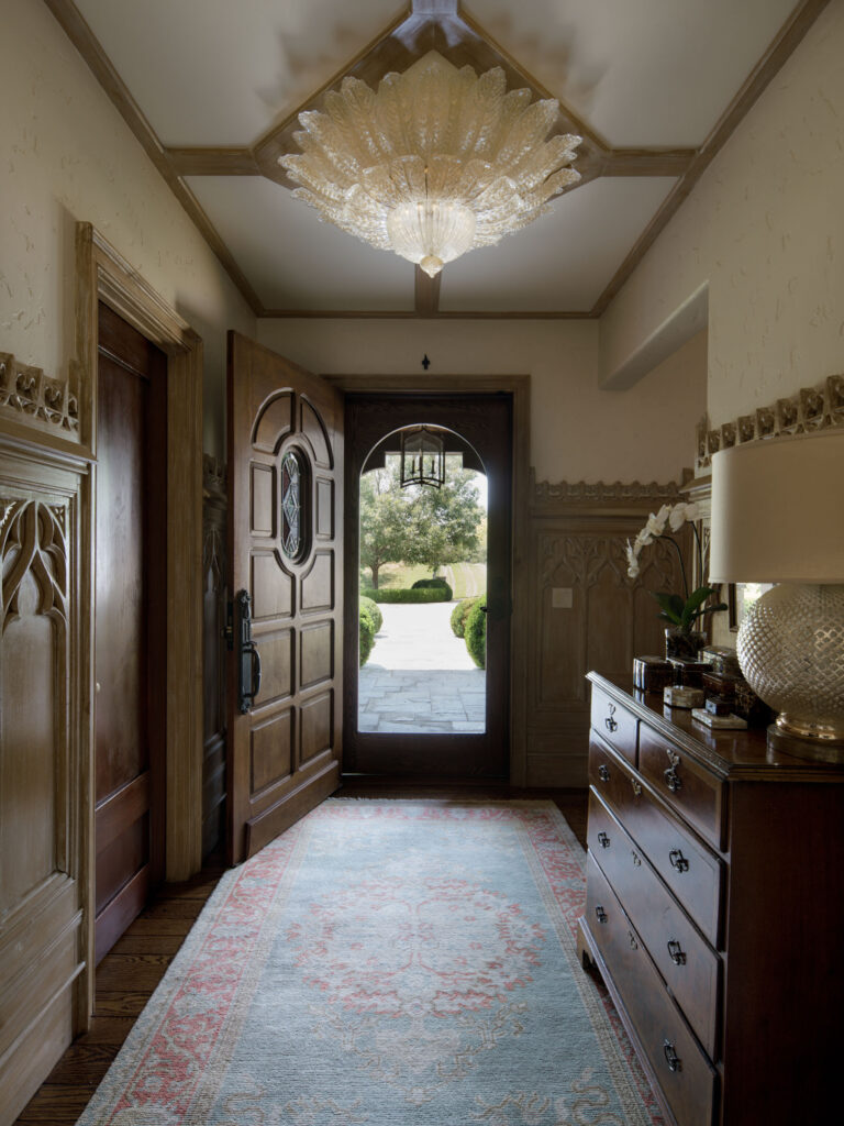 A warmly lit hallway with wood-paneled walls, an ornate wooden door open to a garden view, a vintage rug on a wooden floor, a carved chest with a lamp and flowers, and a decorative glass ceiling light overhead.