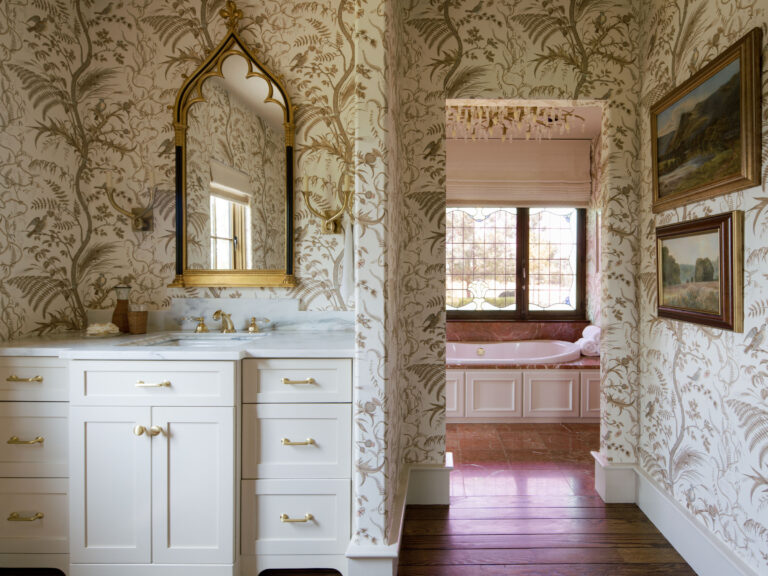 Elegant bathroom with floral-patterned wallpaper, white vanity with gold hardware, marble countertop, and arched mirror. Through an open doorway, a pink bathtub sits beneath large windows. Framed landscape paintings hang on the wall, and wood floors are visible.