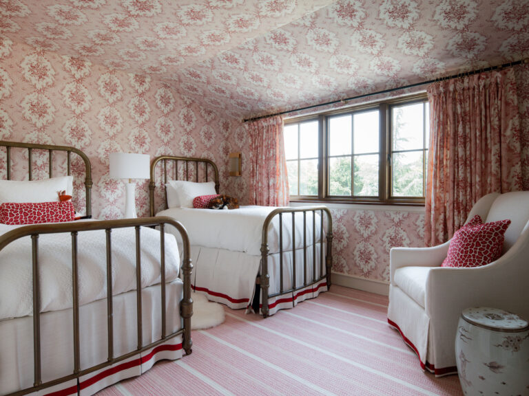 A cozy bedroom with two metal-framed twin beds, white bedding, and red accent pillows reflects the exuberant interior of this French Norman-inspired home, featuring floral wallpaper, pink striped rug, pink curtains, and a window with a green view.