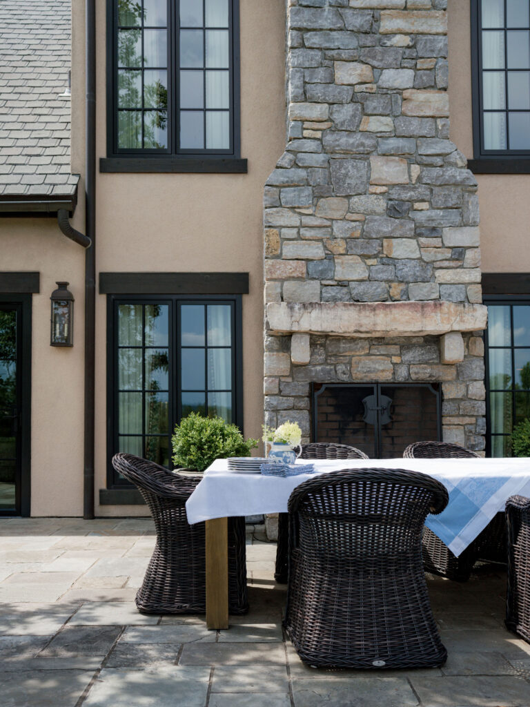 Outdoor patio reflects the exuberant interior of this French Norman-inspired home, with a stone fireplace, beige stucco walls, black-trimmed windows, and a wooden table set with a white and blue cloth. Six dark wicker chairs surround a vase of white flowers.