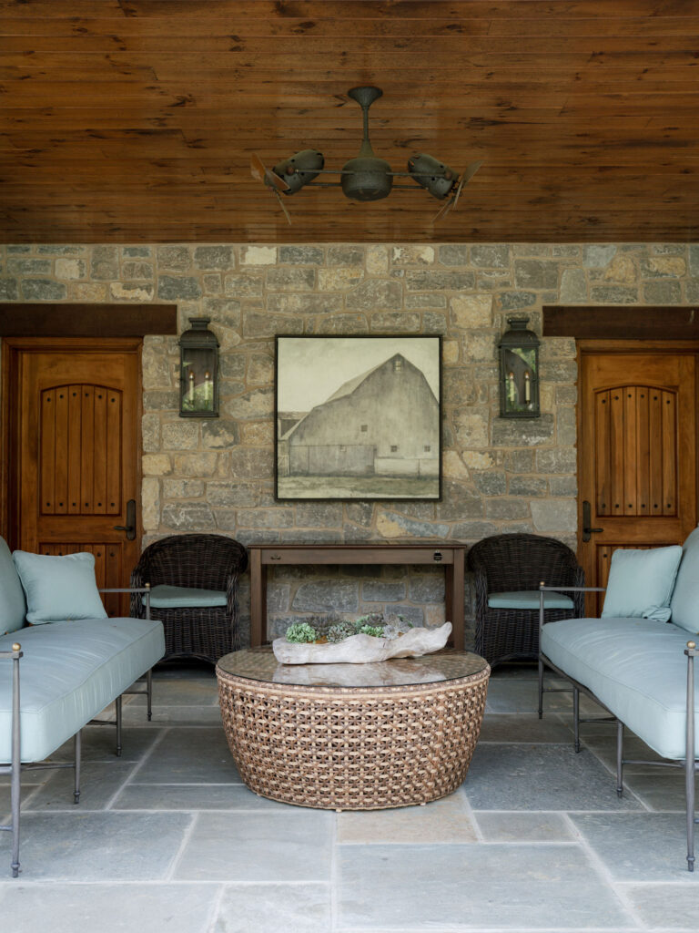 A stone patio in the exuberant interior of this French Norman-inspired home features two light blue cushioned sofas around a round wicker coffee table with a driftwood centerpiece, barn artwork, lanterns, and a wood ceiling with a fan.
