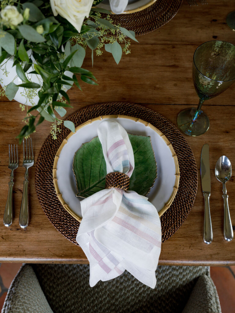 A rustic place setting features a white plate with a green leaf on it, set on a woven charger. A striped cloth napkin, held by a brown napkin ring, rests on the plate. Silver cutlery and a green glass are arranged on a wooden table with greenery above.