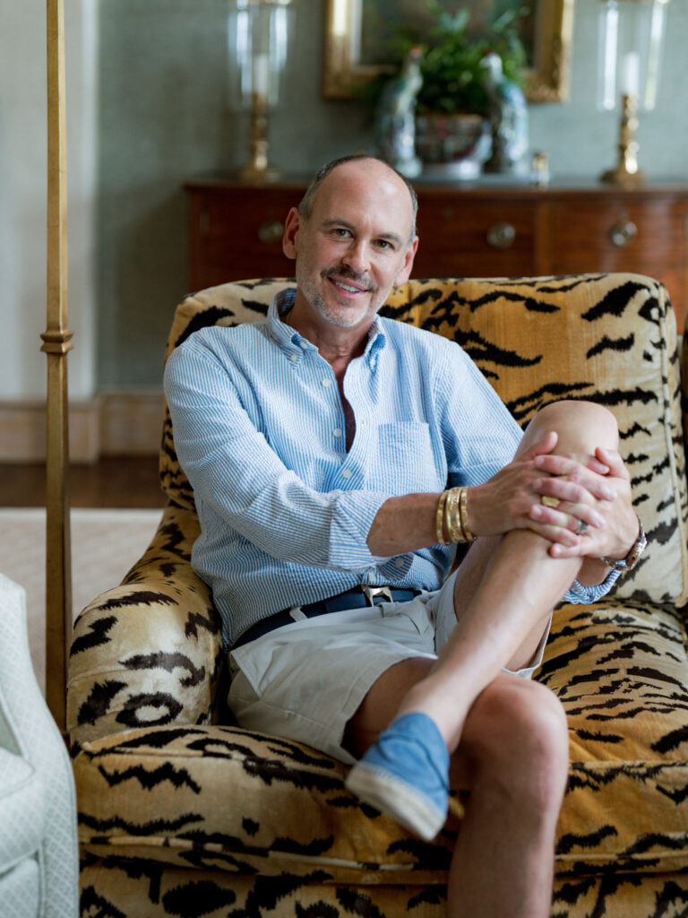 A smiling man sits cross-legged on a tiger-print armchair. He wears a light blue striped shirt, white shorts, blue slip-on shoes, and gold bracelets. Behind him is a wooden sideboard with two candlesticks, decorative items, and a framed mirror.