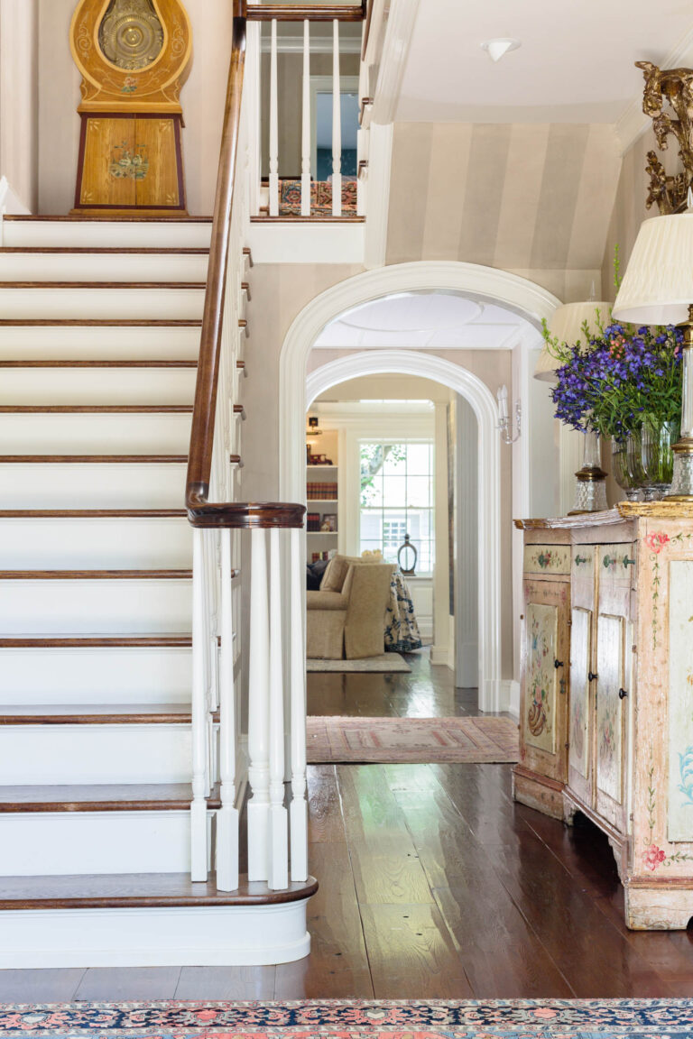 Elegant, sunlit foyer in this 1920s shingled and stone house features wood stairs with white spindles, an ornate painted sideboard with a floral arrangement, arched doorway to a living room with beige sofa, and a grandfather clock at the stair landing.
