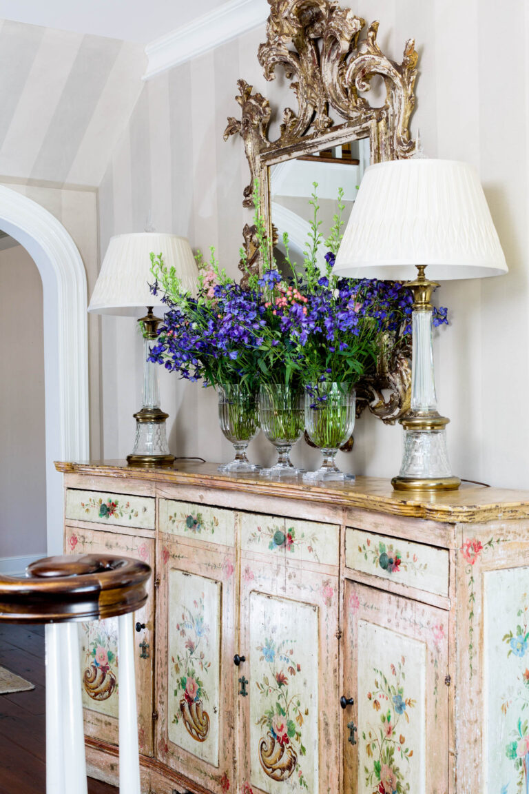 A vintage floral-painted sideboard stands against a striped wall in a 1920s shingled and stone house, topped with ornate table lamps and vases of purple and green blooms. An elaborate gold-framed mirror above reflects the elegant room.