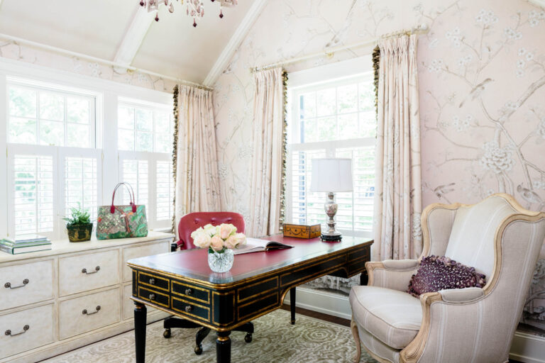 Elegant home office in a 1920s shingled and stone house features floral wallpaper, a large black and gold desk, pink chair, armchair with pillow, books, lamp, vase of roses, and patterned handbag. Sunlight streams through tall windows.