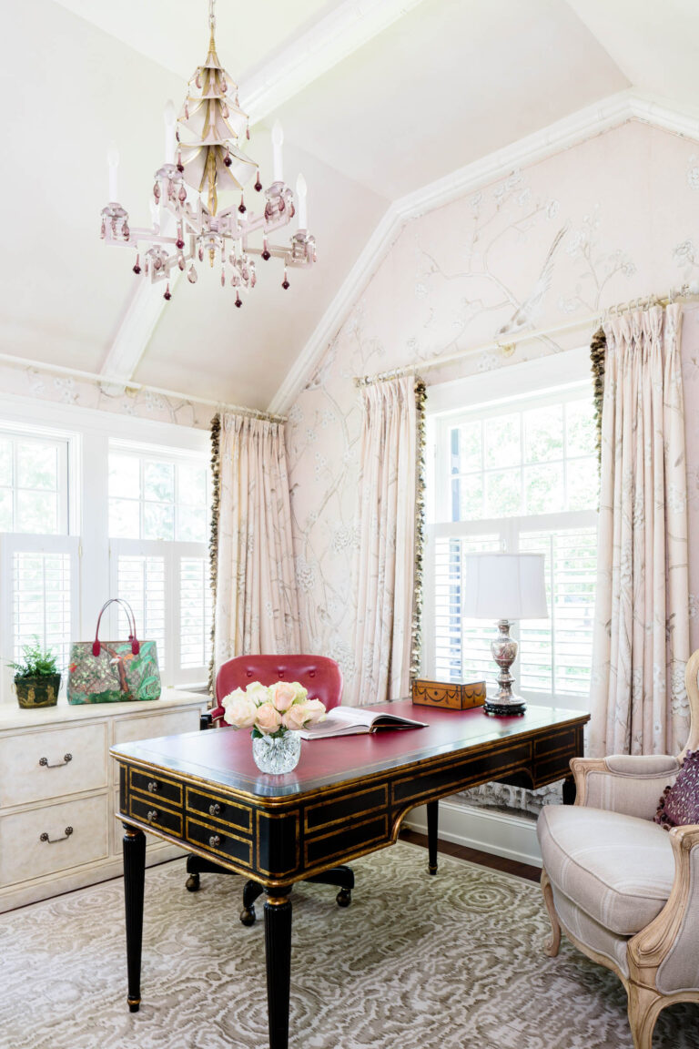 Elegant home office in a 1920s shingled and stone house, with a black and gold desk topped with pink roses, a book, and lamp. Tall windows with light drapes fill the room with sunlight as a chandelier glows overhead above a pink chair.