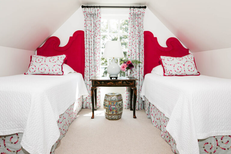 A bright attic bedroom, part of the renovation and expansion of the residence as a weekend destination, features twin beds with bold pink headboards, white bedding with pink accents, and a charming window nook with patterned curtains and fresh flowers.