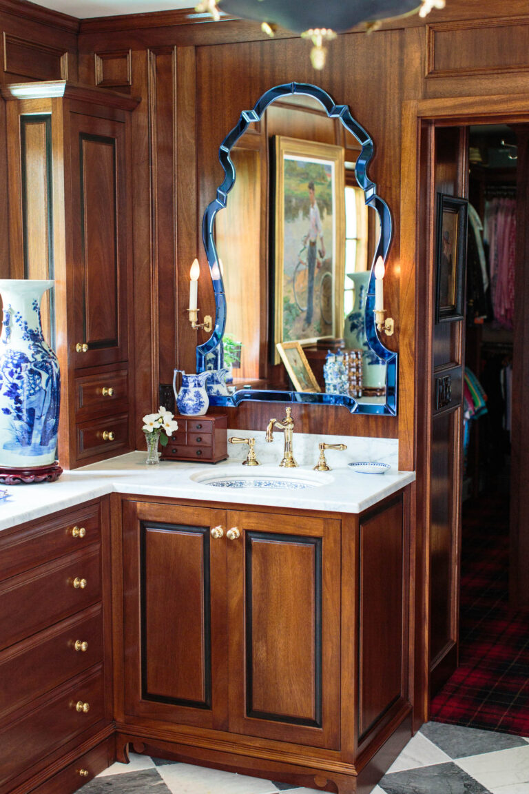 A luxurious bathroom with rich wood cabinetry, gold fixtures, and a blue-trimmed ornate mirror exudes the exuberant interior of this French Norman-inspired home. Blue-and-white vase, flowers, and lit sconces decorate the space; a closet is visible behind.