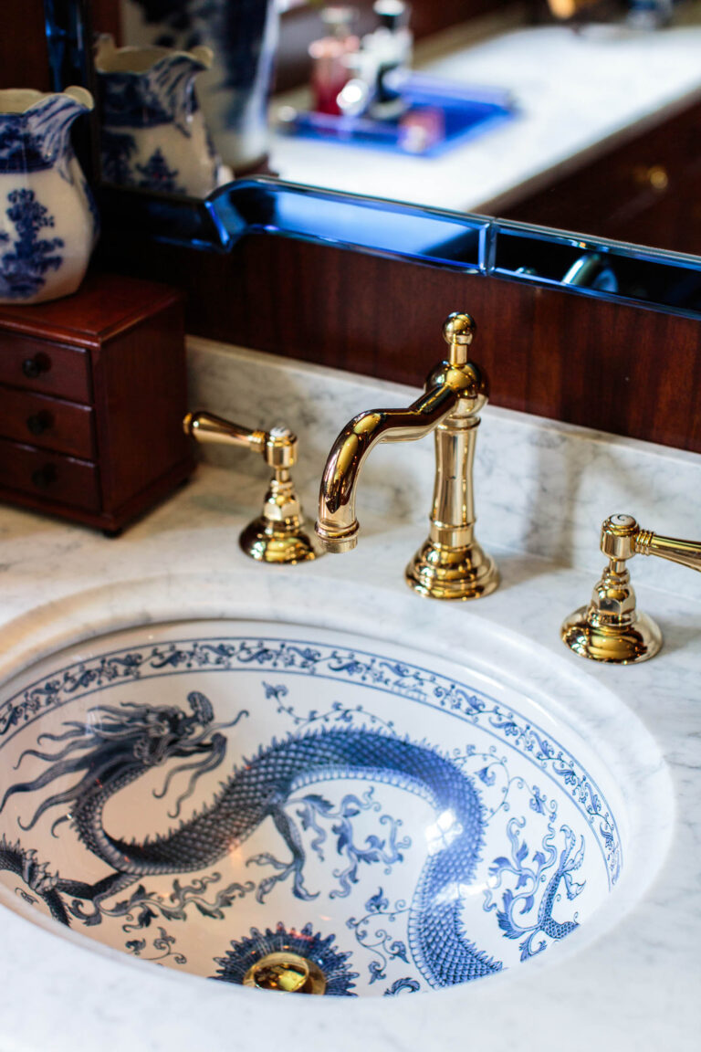 A white marble bathroom sink with an intricate blue dragon design and shiny gold faucets sits amid the exuberant interior of this French Norman-inspired home. A blue and white vase, small wooden chest, and mirror complete the elegant countertop scene.