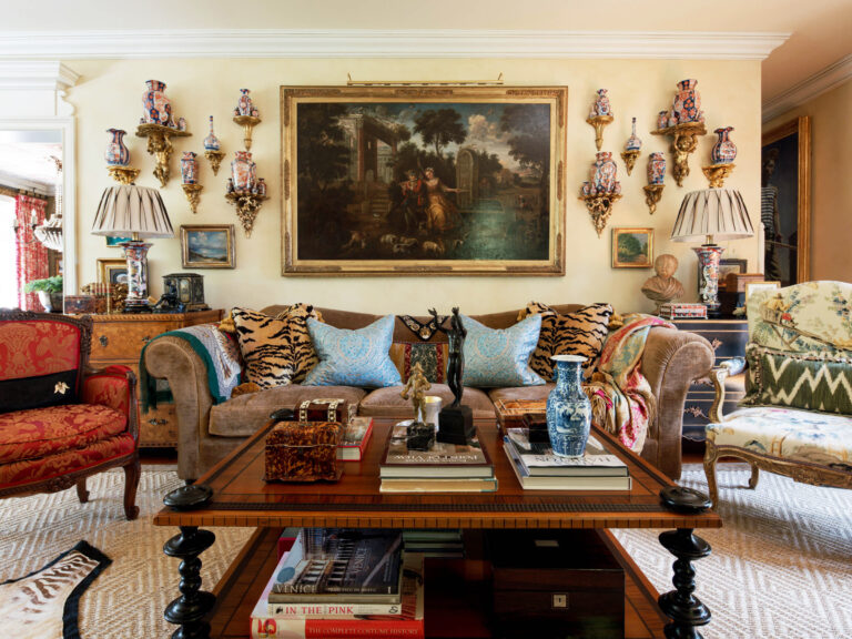 A richly decorated living room with an ornate sofa featuring patterned and tiger-print pillows. The wall has a large framed painting and many porcelain vases. A wooden coffee table holds books, decorative objects, and vases. The room has eclectic, elegant decor.