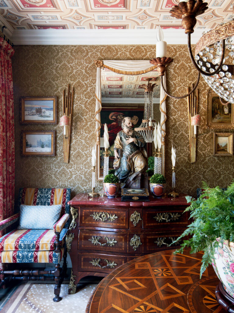 An ornate, traditional room with patterned walls, a mirror above a wood chest, a religious statue, candelabras, framed art, a striped armchair with cushions, crystal chandelier, potted plants, and a table with intricate wood inlay. The ceiling is also decorated.