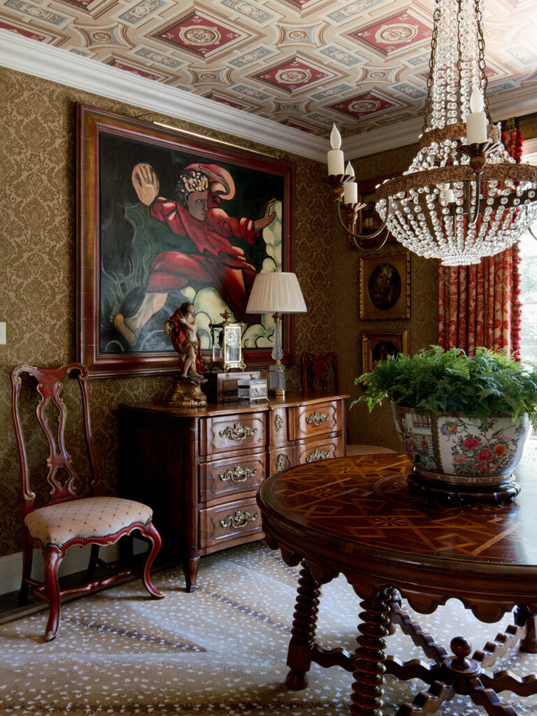 A richly decorated room features ornate wallpaper, a patterned ceiling, and a crystal chandelier. There is a wooden chest with a lamp, figurines, and a dramatic painting above. A carved chair and a round table with a large potted plant complete the elegant scene.