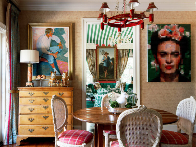 Eclectic dining room with a round wooden table, plaid cushioned chairs, and a gold chest of drawers. Colorful portraits, including a pixelated Frida Kahlo, hang on the walls. A red chandelier hangs above. The adjoining room is visible through decorative curtains.