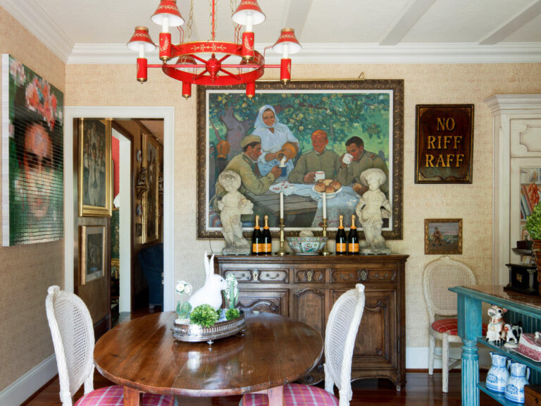A dining room with a round wooden table and four plaid chairs, a red chandelier above, and a large painting of people eating on the back wall. Theres an ornate sideboard with champagne bottles, two busts, and a “NO RIFF RAFF” sign on the right wall.