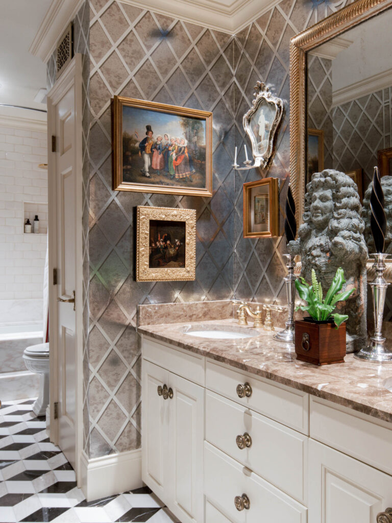 Elegant bathroom with diamond-patterned wallpaper, marble countertop, and white cabinets. Classic framed art and a decorative bust are displayed above the sink. Gold fixtures, a small potted plant, and geometric tile flooring add to the sophisticated decor.