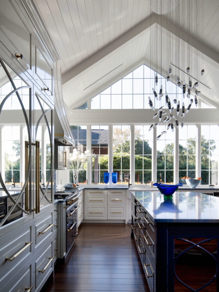 A spacious, modern kitchen with a vaulted, white-paneled ceiling and floor-to-ceiling windows showcases the exuberant interior of this French Norman-inspired home, featuring glossy white cabinets, a navy island, wood floors, and a sculptural glass chandelier.