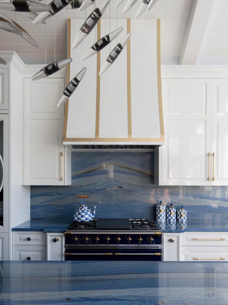 A modern kitchen with white cabinets, gold accents, and a striking blue marble backsplash reflects the exuberant interior of this French Norman-inspired home. A navy stove sits below a white and gold range hood, with blue-and-white checkered canisters decorating the counter.