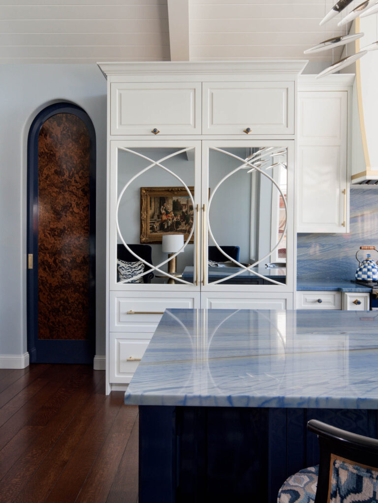 A modern kitchen in the exuberant interior of this French Norman-inspired home features glossy blue marble countertops, white cabinetry with gold handles, mirrored cabinet doors with geometric designs, and rich dark wood floors.