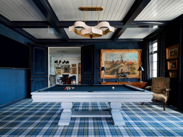 A stylish game room with a plaid carpet, navy blue paneled walls, and a white billiard table set for play, showcasing the exuberant interior of this French Norman-inspired home. A gold-framed painting, antique chair, and modern chandelier complete the look.